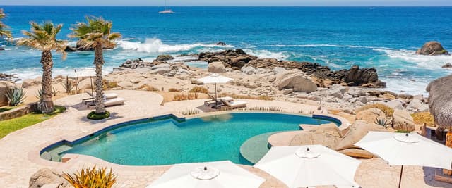 Luxury villa with infinity pool, lounge chairs, and illuminated terrace overlooking coastal bay at dusk