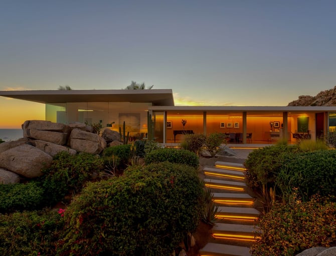Modern single-story house with glass walls illuminated at dusk, surrounded by desert landscaping, boulders, and lit pathway overlooking ocean at sunset.